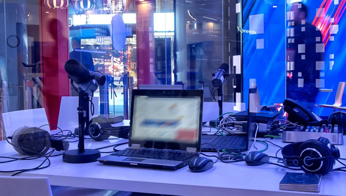 Podcast recording studio with a table, microphones, laptop, headphones, and audio equipment, in a modern, glass-walled setting.