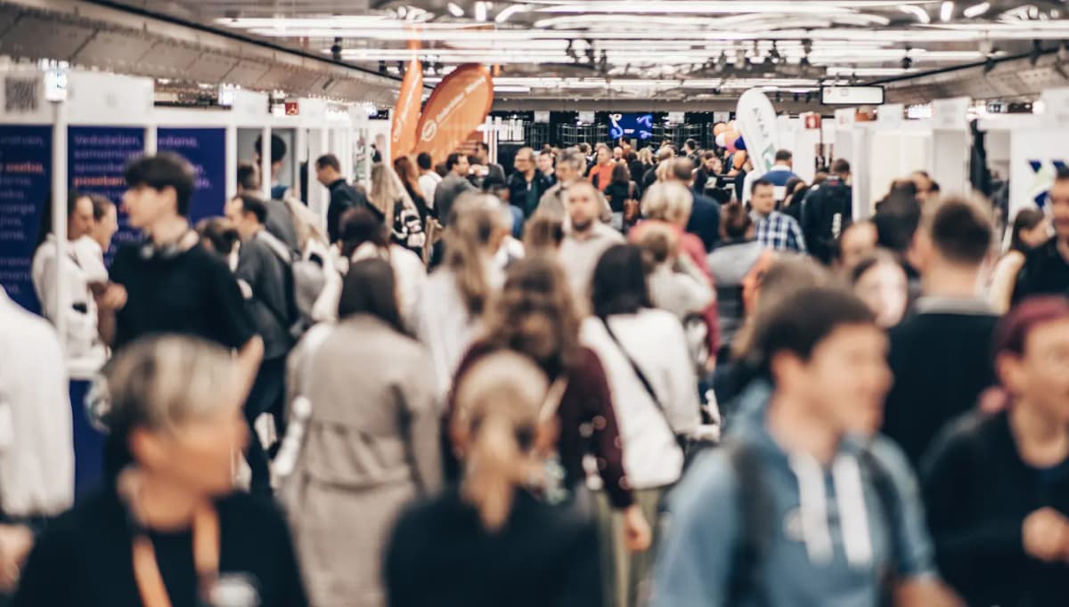 A crowded convention hall with people walking between booths, banners, and displays in a bustling atmosphere.