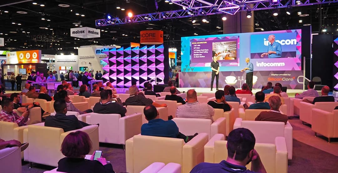 Audience seated on white couches watching a presentation on stage at a tech conference. The backdrop features geometric designs and colorful lighting.