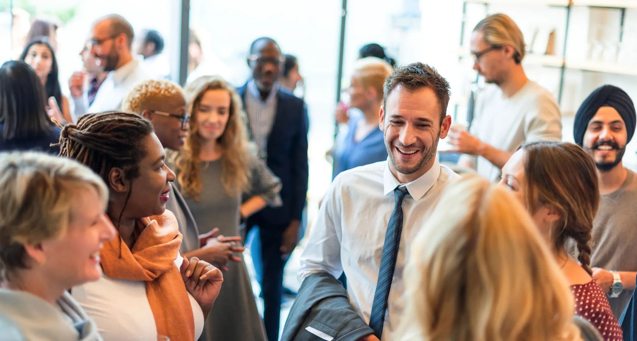 A group of people engaged in lively conversation at a social gathering, with one person in a tie smiling and others chatting around.