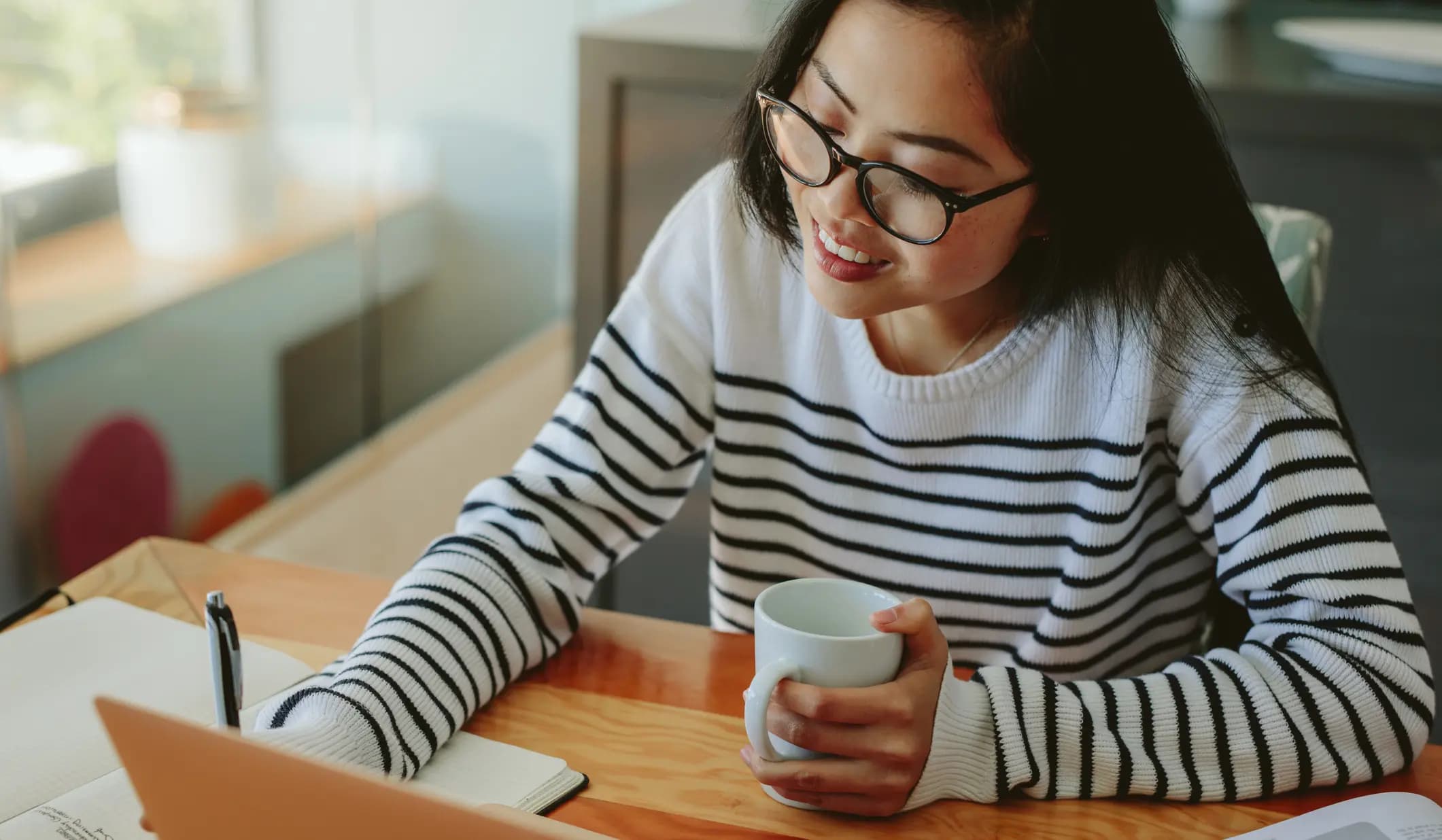 A woman in glasses and a striped sweater writes in a notebook while holding a mug, sitting at a wooden table with a laptop in front of her.