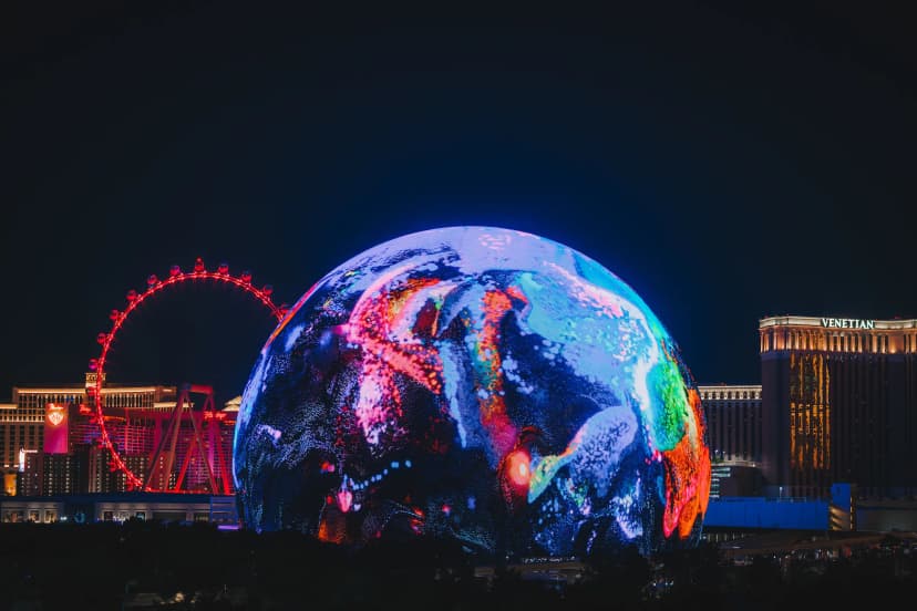 A colorful, illuminated spherical structure with vibrant patterns against a night sky, with a red Ferris wheel and lit buildings in the background.
