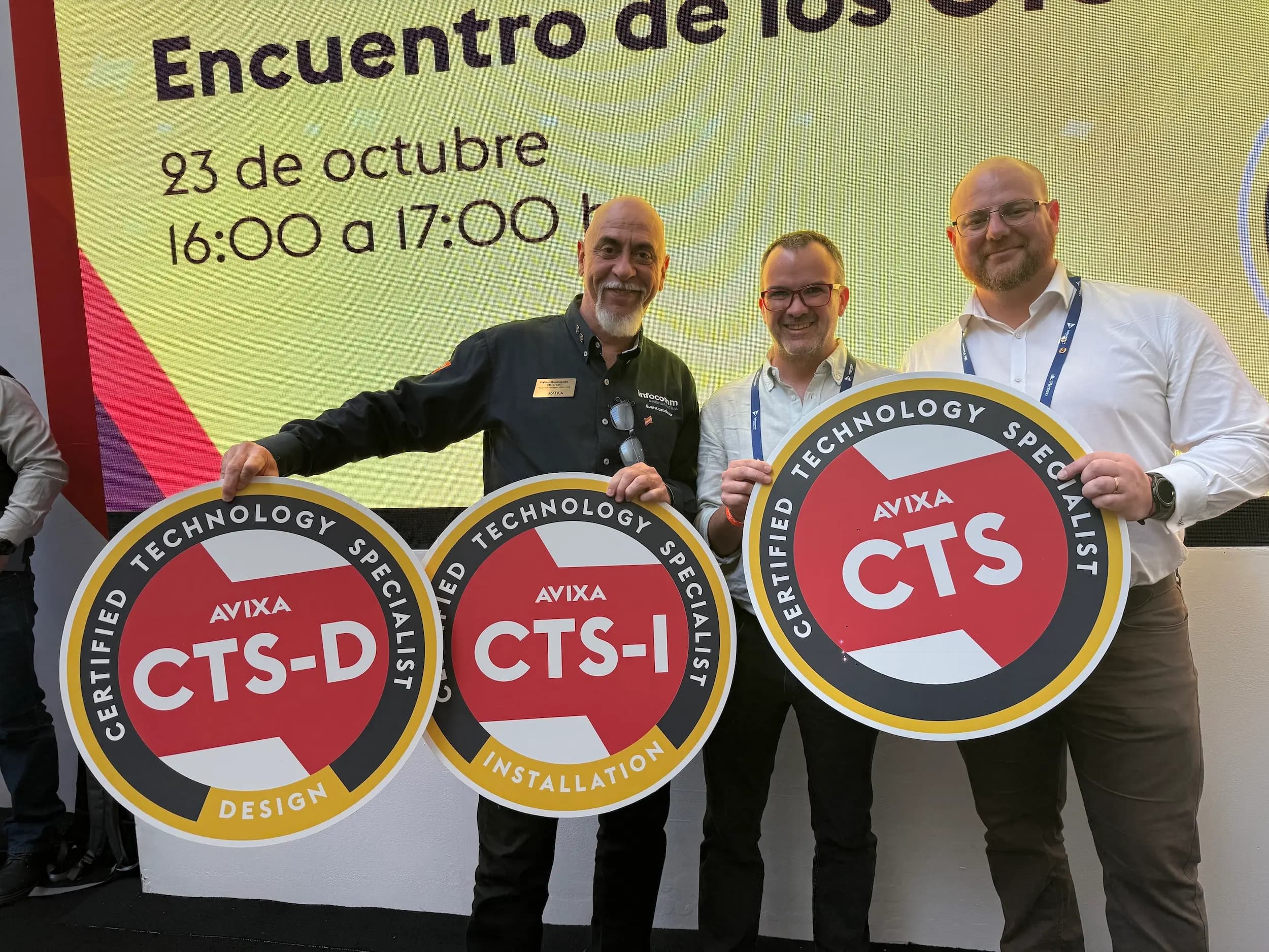 Three men holding AVIXA Certified Technology Specialist signs at an event. The background displays event details in Spanish.