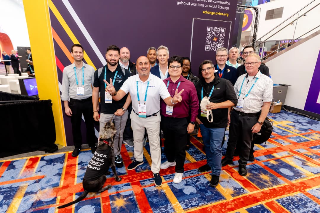 A group of people, wearing conference badges, pose together on a colorful carpet at a conference booth, with a dog wearing a promotional shirt.