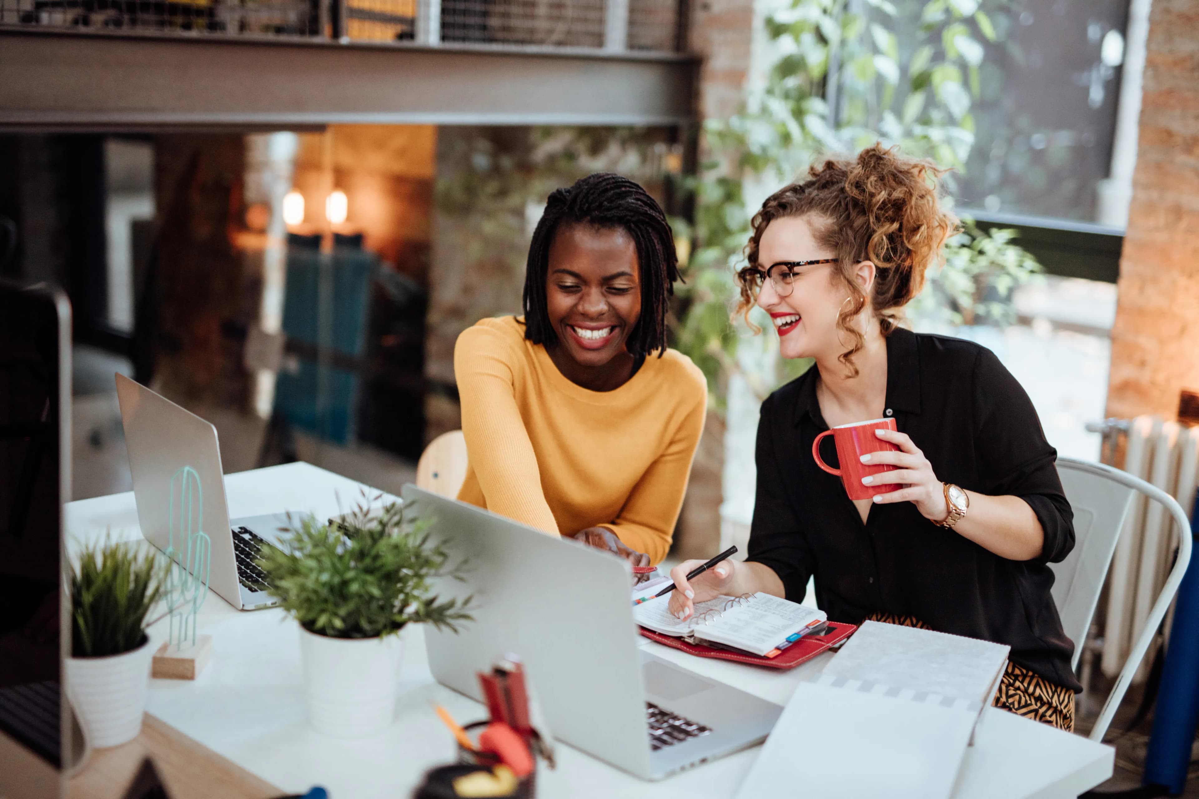Two women smiling and collaborating at a desk with laptops, a red mug, and notebooks. Bright office space with plants.