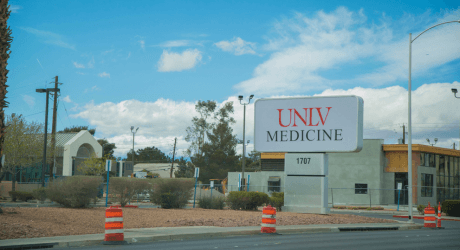 Sign reading "UNLV Medicine" beside a road with orange barrels, set against a partly cloudy sky and surrounding buildings and trees.