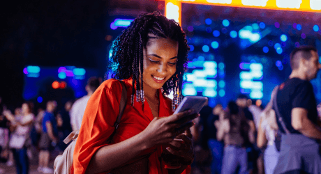 A woman in a bright orange top smiles while looking at her phone at a lively outdoor event with colorful lights in the background.