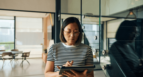 A woman with glasses and a striped shirt uses a tablet in a modern office with large windows and minimalistic decor.