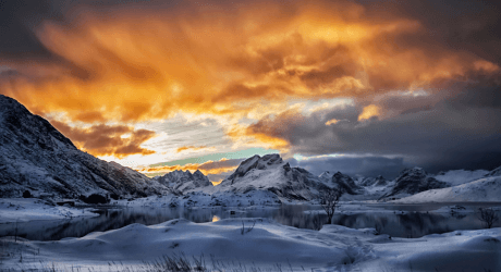 Snow-covered mountains and a calm lake under a dramatic, fiery sunset sky with orange and gray clouds.
