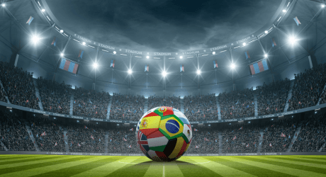 Soccer ball with various national flags on the grass in a brightly lit stadium with a cheering crowd.
