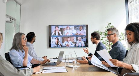 People in a conference room are having a meeting with colleagues on a screen. Papers and laptops are on the table.