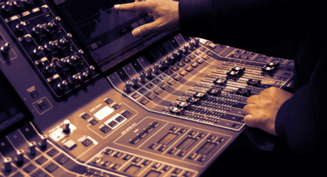 Hands adjusting audio mixing console sliders and knobs, with lit buttons and a screen displaying controls, in a dimly lit studio.