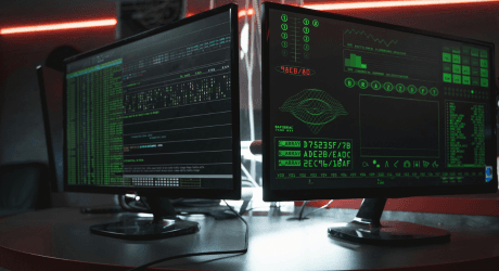 Dual-monitor setup displaying green-on-black code, system graphs, and data streams in a dark room, illustrating a hacker-style cyberwar operation or cybersecurity breach analysis.