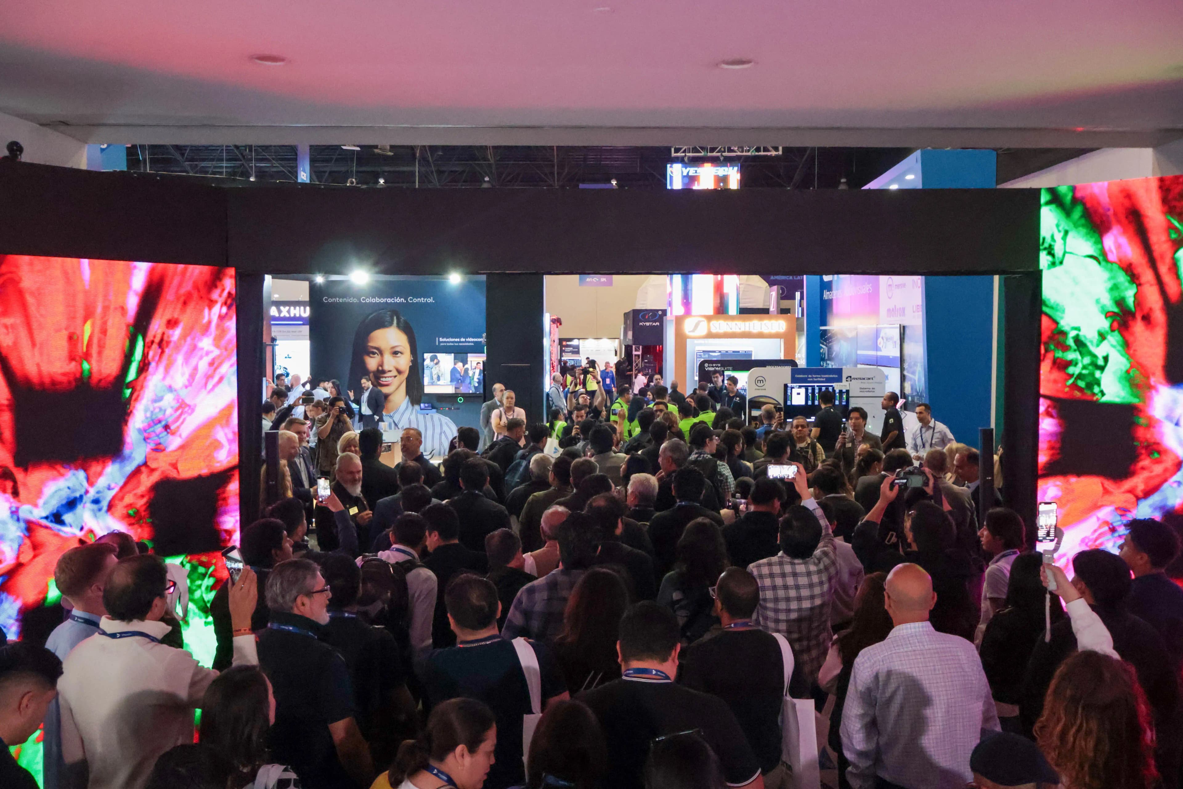 A crowded tech expo with attendees gathered around colorful digital displays and booths.