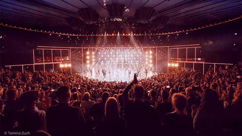 A large audience in a theater applauding performers on a brightly lit stage with spotlights and decorative vertical lights.