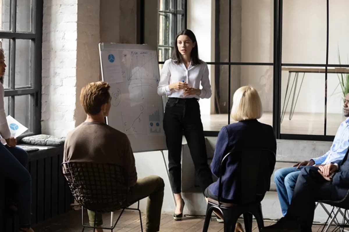 A woman presents to a group seated in a circle, using a flip chart in a modern office with large windows.