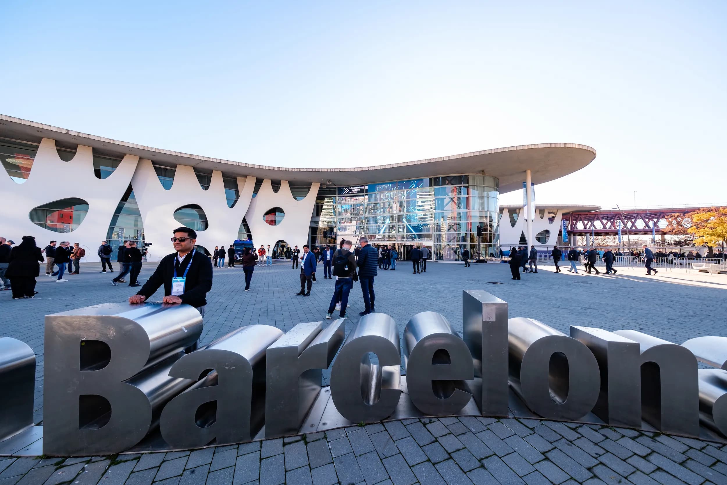 People gather outside a modern building with unique white facade. Large "Barcelona" letters in the foreground.