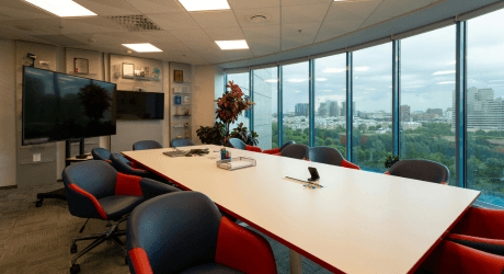 Modern conference room with a long white table, blue and red chairs, large TV screen, glass wall, and cityscape view.