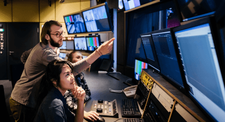 People working at a control room desk, surrounded by monitors and broadcasting equipment, focusing on video editing or production tasks.