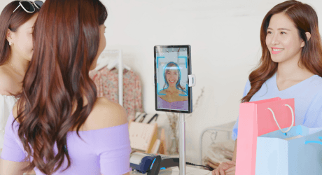 Two women at a store counter, one using facial recognition on a tablet for payment, with shopping bags visible.