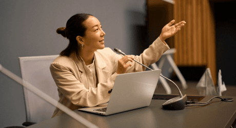 Woman speaking at a conference table, gesturing with her hand, using a laptop and microphone.