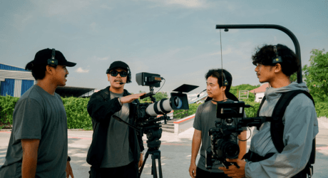 Film crew discussing a scene outdoors, with one person gesturing near a camera on a tripod, wearing headsets and casual clothing.