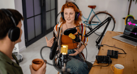 Two people recording a podcast in a casual setting, seated at a table with microphones and headphones, laptop showing audio waves.