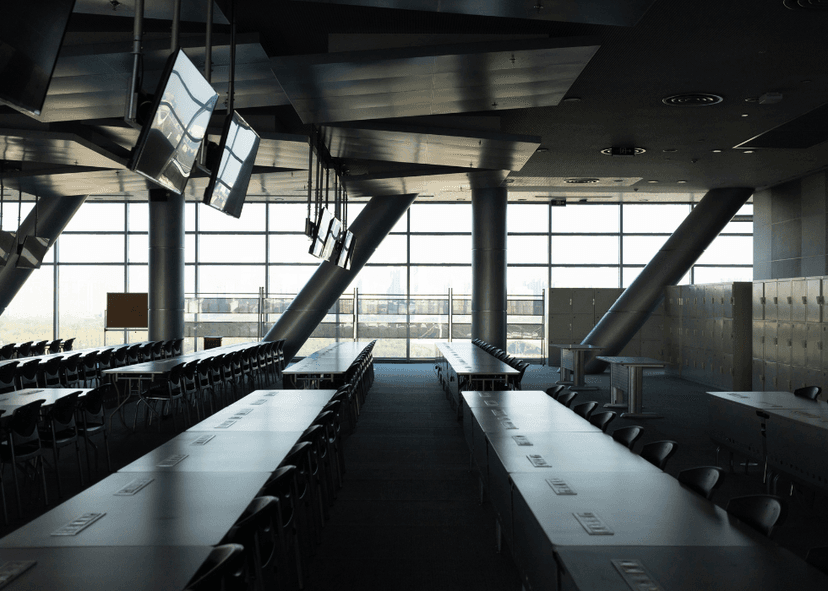 Modern classroom with long tables and chairs in rows, large windows along one side letting in daylight, clean minimalist interior, set up for group learning and presentations.